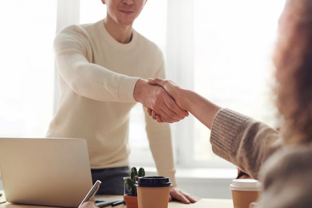 two people shaking hands over a desk with an open laptop on it